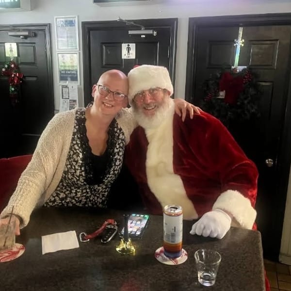 A smiling woman with a drink poses with Santa Claus at a bar table.
