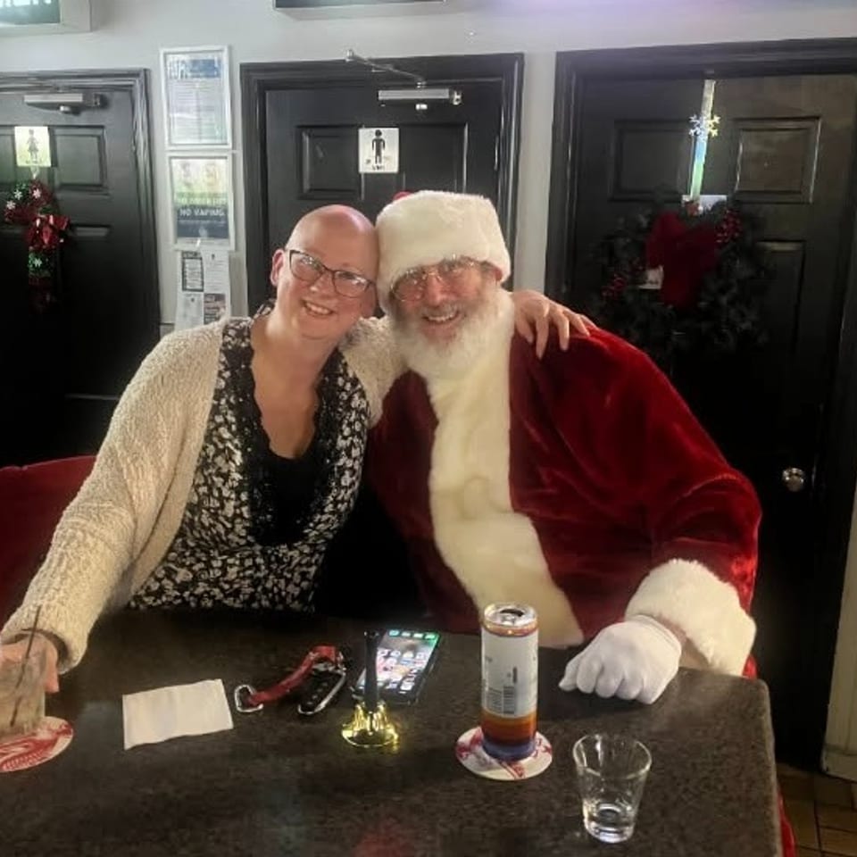 A smiling woman with a drink poses with Santa Claus at a bar table.