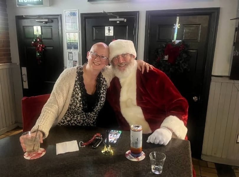 A smiling woman and Santa Claus pose together at a bar table with drinks.