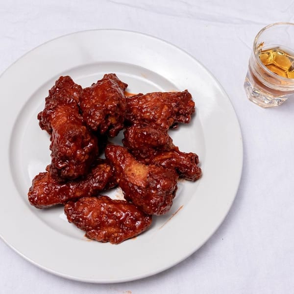 A white plate holds several saucy chicken wings next to a glass of amber liquid.
