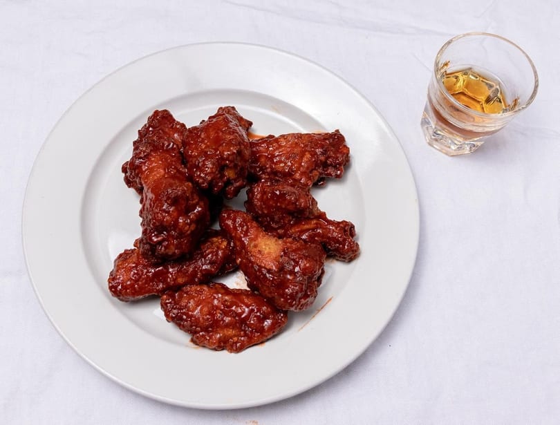 A white plate holds several saucy chicken wings next to a glass of amber liquid.