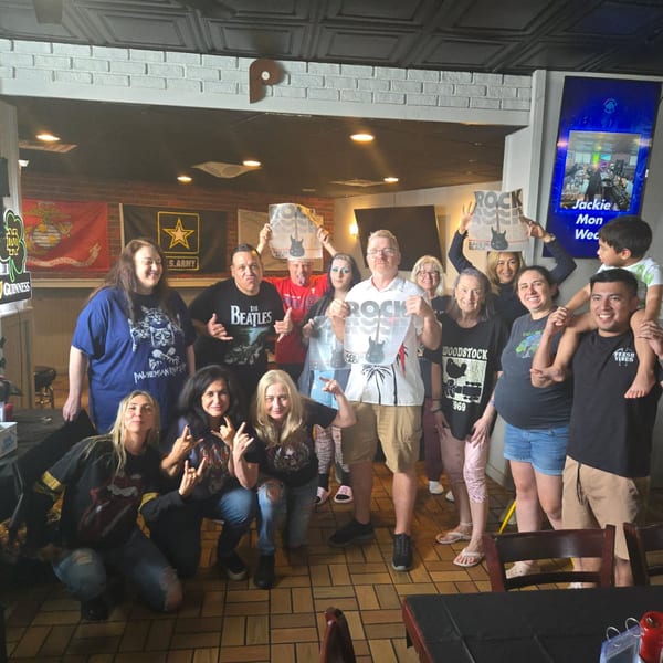 A diverse group of people in rock band shirts pose together in a casual restaurant setting.