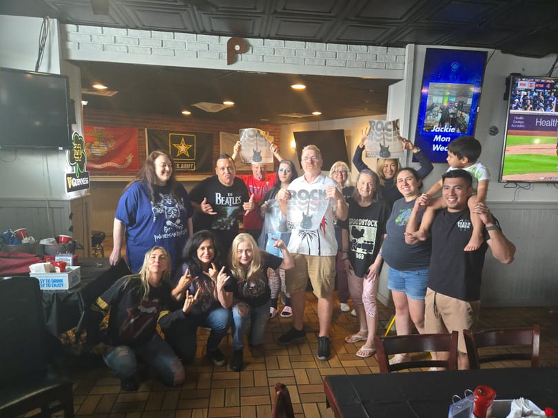 A diverse group of people in rock band shirts pose together in a casual restaurant setting.