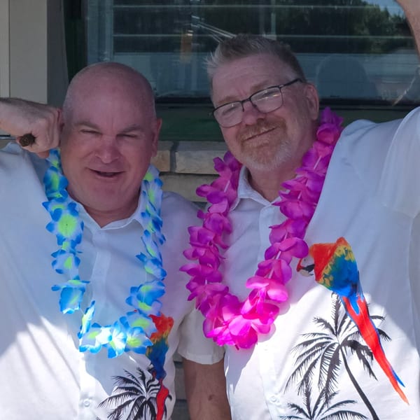 Two smiling men in white Hawaiian shirts and leis, one blue and one pink, posing outdoors.