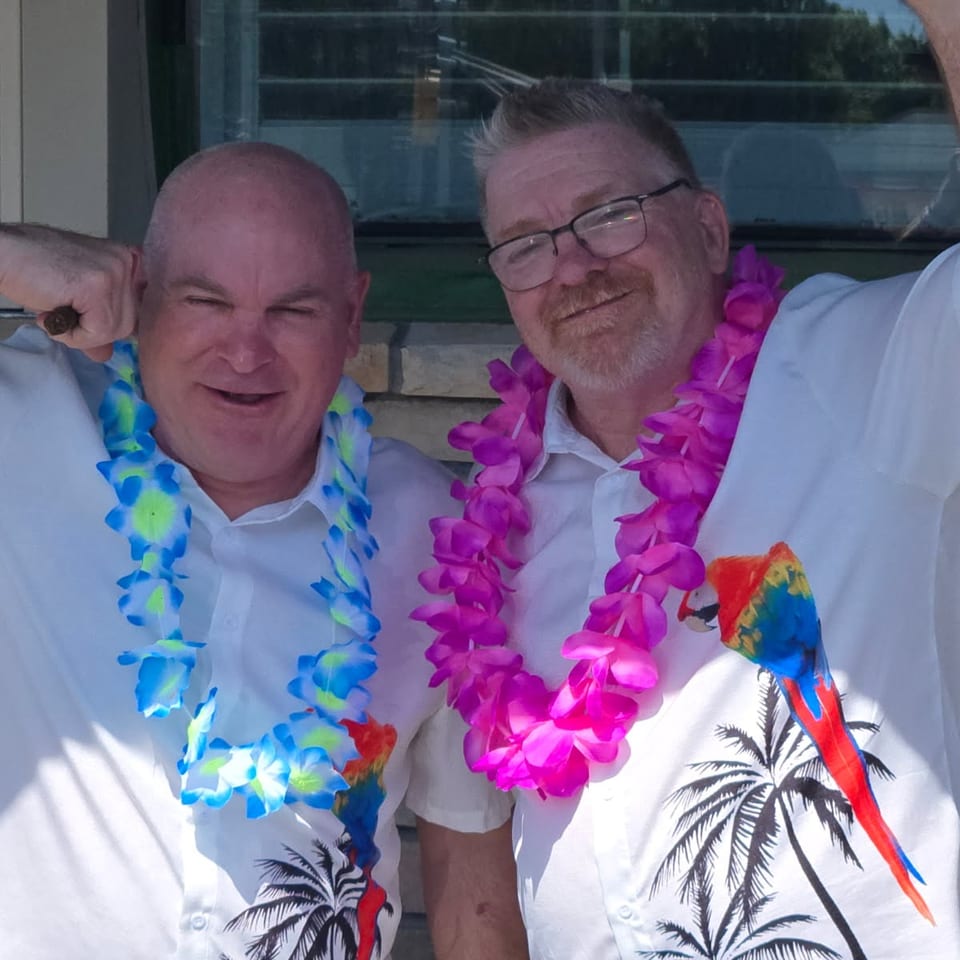 Two smiling men in white Hawaiian shirts and leis, one blue and one pink, posing outdoors.