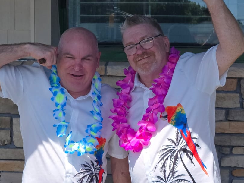 Two smiling men in white Hawaiian shirts and leis, one blue and one pink, posing outdoors.
