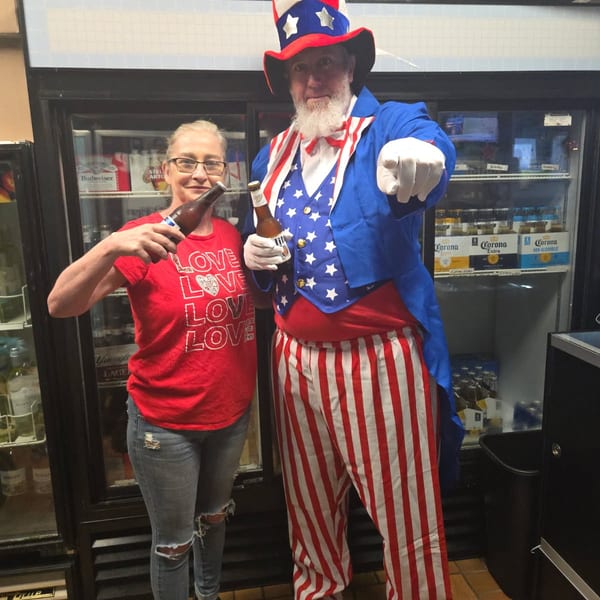 A woman and a man dressed as Uncle Sam hold beer bottles in front of refrigerators.