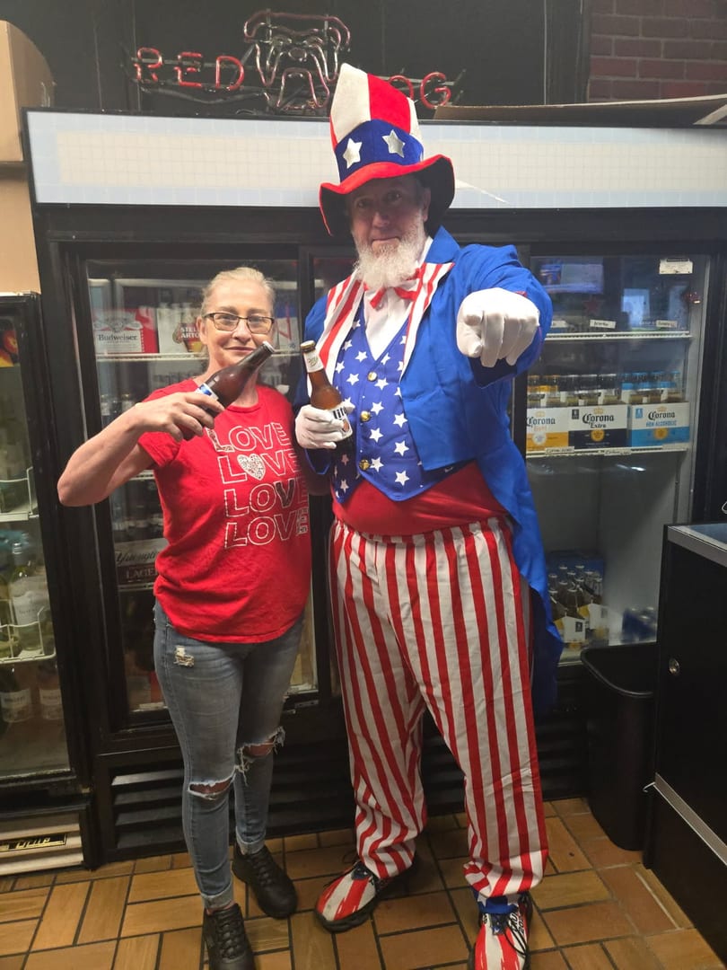 A woman and a man dressed as Uncle Sam hold beer bottles in front of refrigerators.