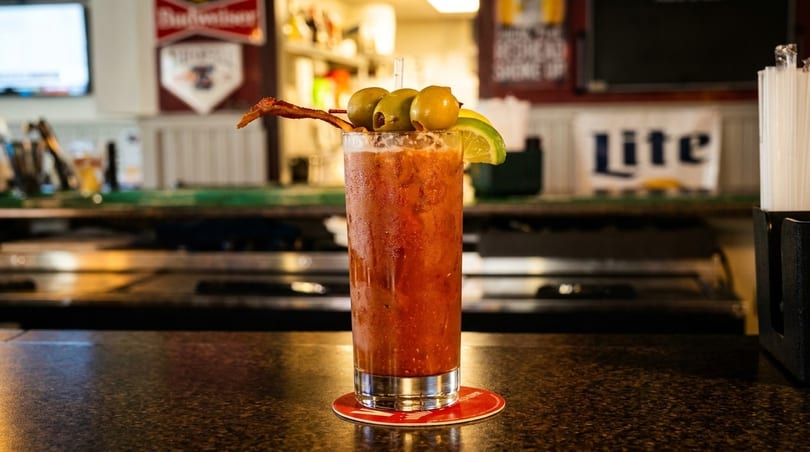 A Bloody Mary cocktail garnished with olives, lime, and bacon, on a bar counter.