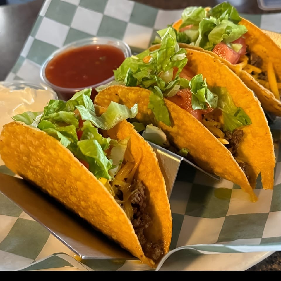 Four crispy tacos filled with ground beef, cheese, lettuce, and tomato, with salsa and sour cream.