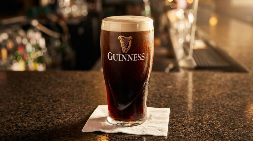 A pint of Guinness beer with a creamy head sits on a napkin on a dark bar.